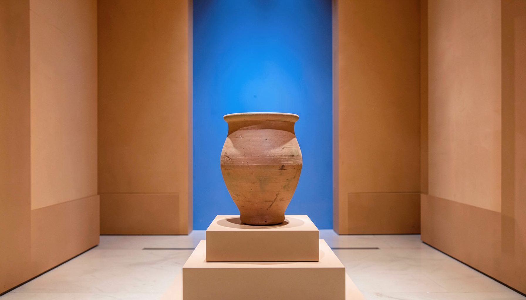 A symmetrical view of an ancient clay vessel on a pedestal, museum gallery setting, soft ochre and terracotta hues