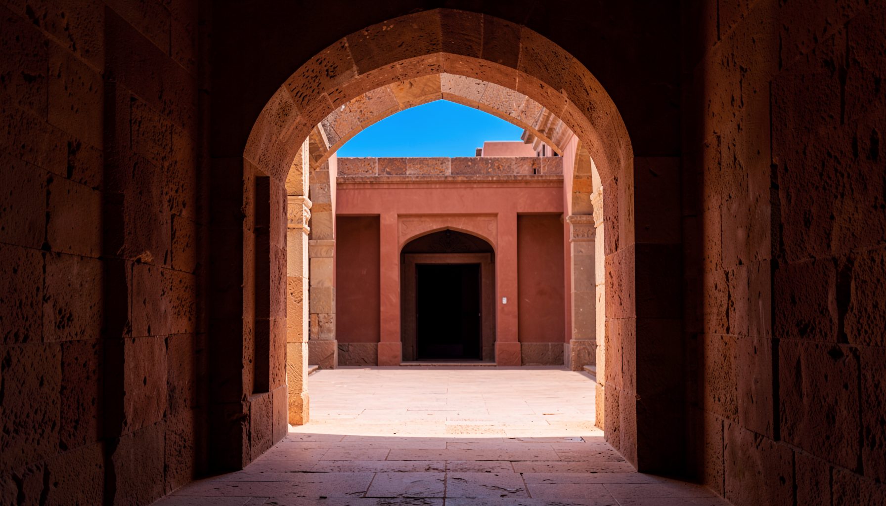 Warmly lit stone archway framing a distant courtyard, deep terracotta and sand tones, tranquil ancient atmosphere