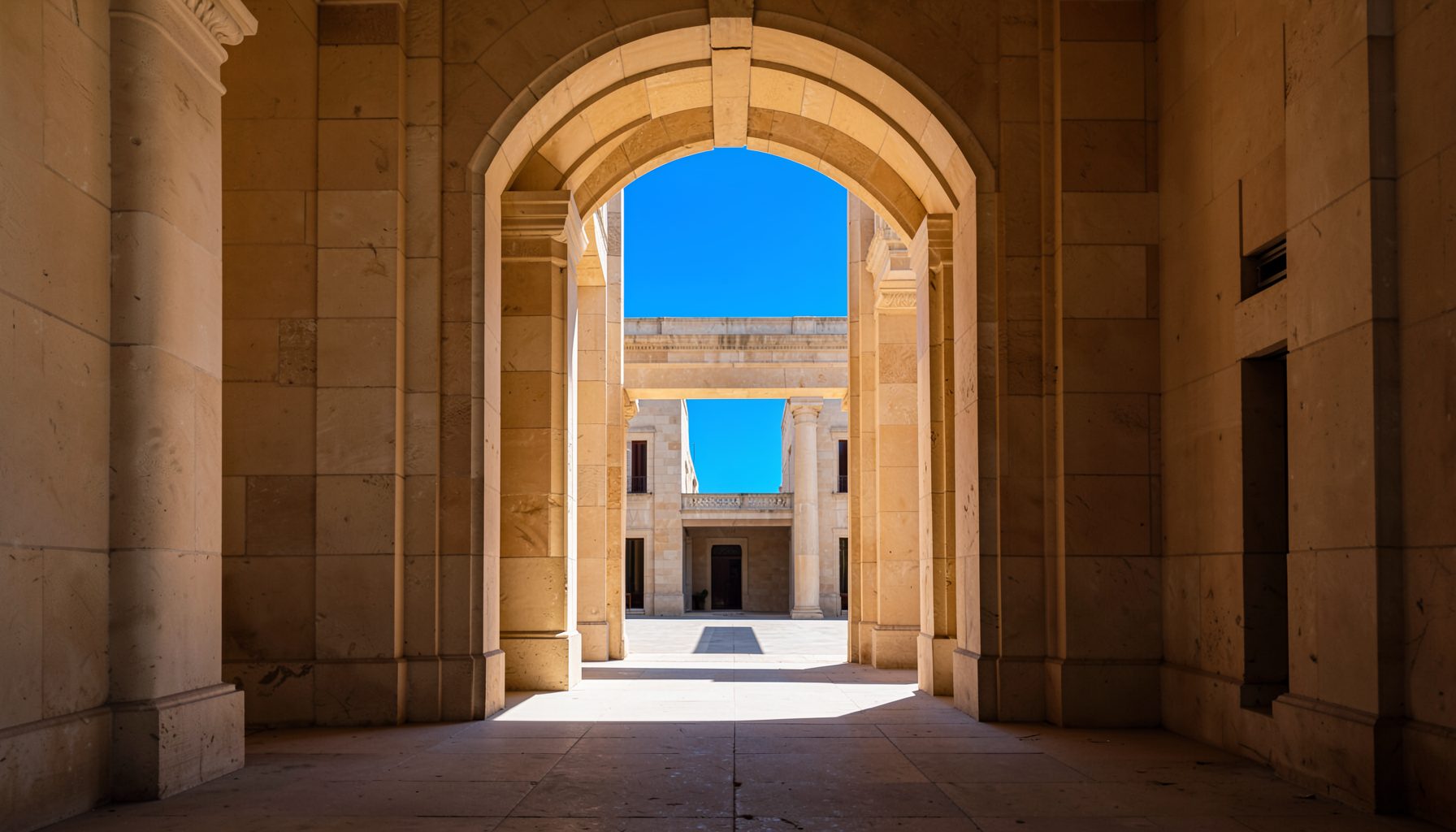 A grand stone archway in warm beige and ochre tones, framing a sunlit courtyard beyond, with strong symmetrical lines and deep shadows