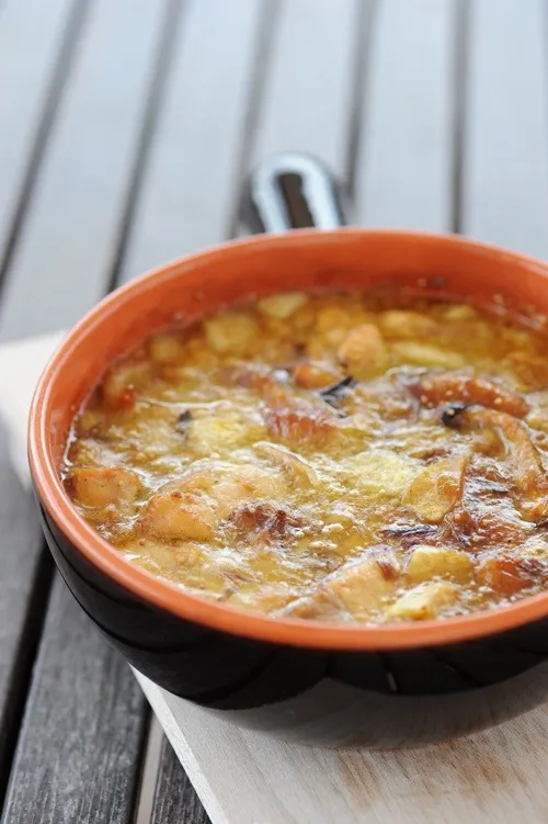 Close-up of a rustic bowl filled with a baked cheesy potato and meat dish on a wooden table; warm orange and brown tones