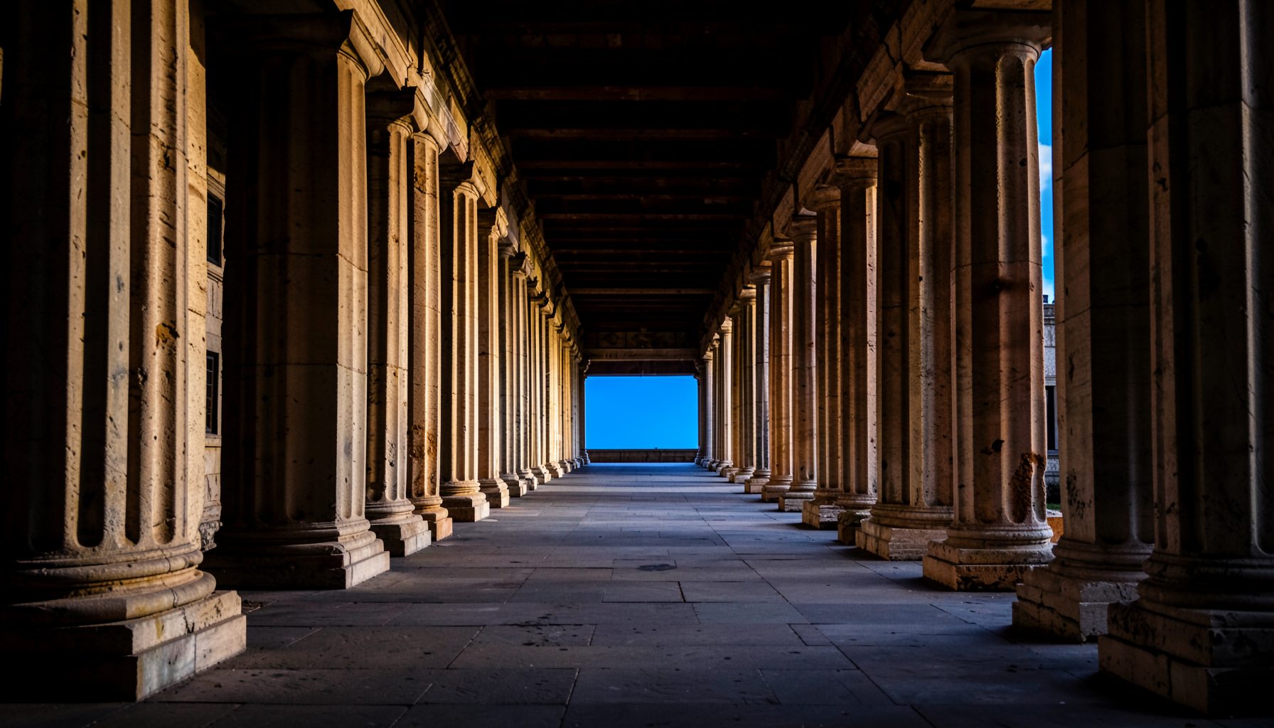 A grand classical colonnade receding in strong perspective, warm stone tones against deep shadow, conveying the weight of ancient history