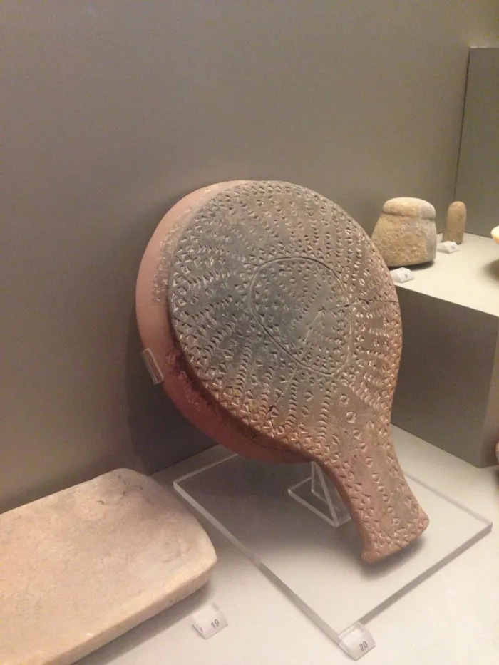 Museum display of an ancient textured ceramic artifact shaped like a paddle, beige and gray tones, with other artifacts nearby
